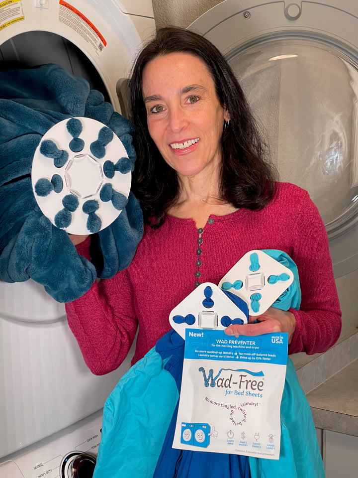 Wad-Free Inventor Cyndi Bray stands by an open dryer door with white round product Wad-Free for Blankets & Duvet Covers attached to a dark teal blanket, and two white square Wad-Free for Bed Sheets attached to teal and blue bed sheets