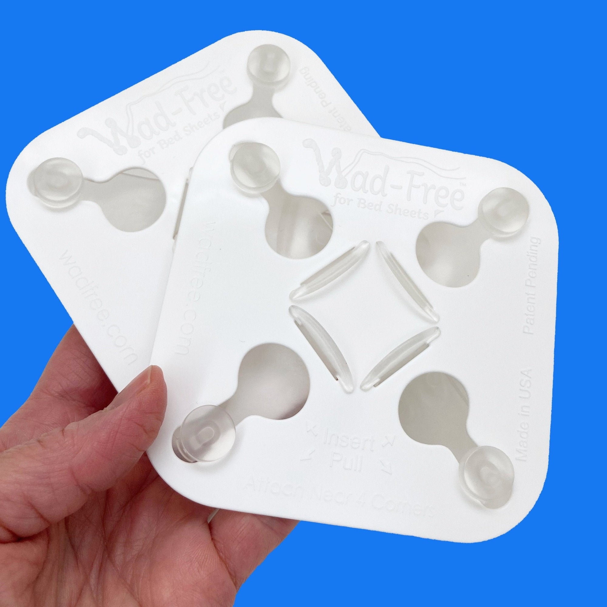 A hand holds two white Wad-Free squares. The product has four tabs with buttons in a keyhole shape and has the logo embossed on it