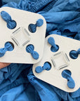 A hand holds a white Wad-Free square attached at four points to a blue fitted sheet, Next to it is a Wad-Free on a flat sheet