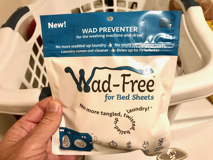 Hand holding a blue and white package of Wad-Free for Bed Sheets with a white laundry basket in the background