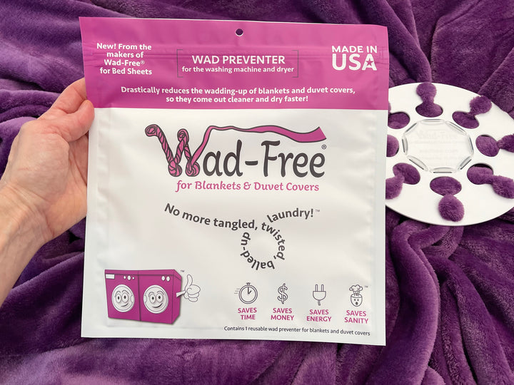 Hand holding an orchid and white package of Wad-Free for Blankets and Duvet Covers with the white round product attached to a purple blanket behind it