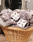 Two grey sheets attached to two white square Wad-Frees are sitting in a wicker laundry basket next to an open clothes dryer door