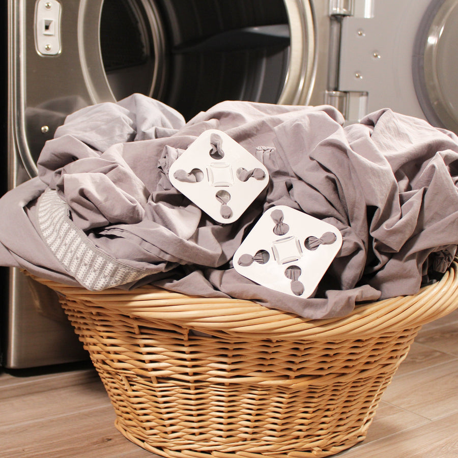 Two grey sheets attached to two white square Wad-Frees are sitting in a wicker laundry basket next to an open clothes dryer door