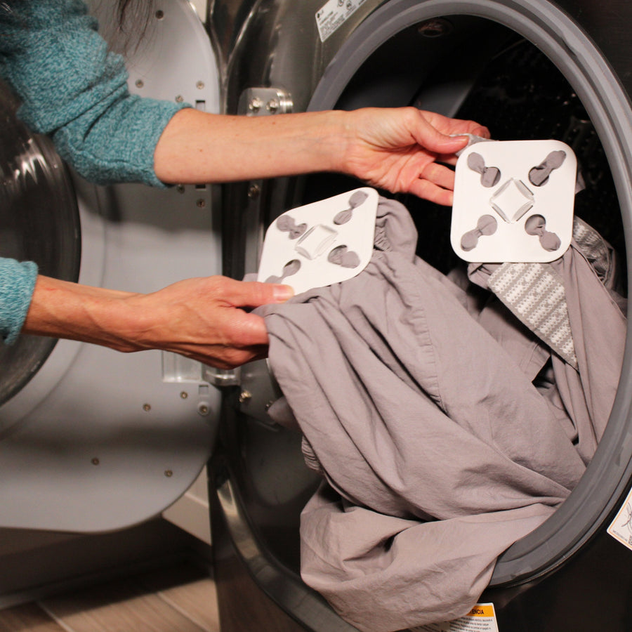 Two hands are shown reaching into an open dryer taking two grey sheets out with each attached to white Wad-Frees