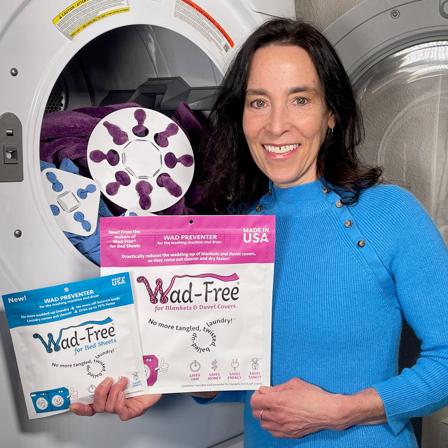 Wad-Free inventor Cyndi Bray in a blue dress holds packaging of Wad-Free for Bed Sheets and  Wad-Free for Blankets and Duvet Covers. The packaging is blue and orchid. One white square is seen in the open clothes dryer attached to a blue sheet and one white round Wad-Free is shown attached to a purple blanket in the open clothes dryer 